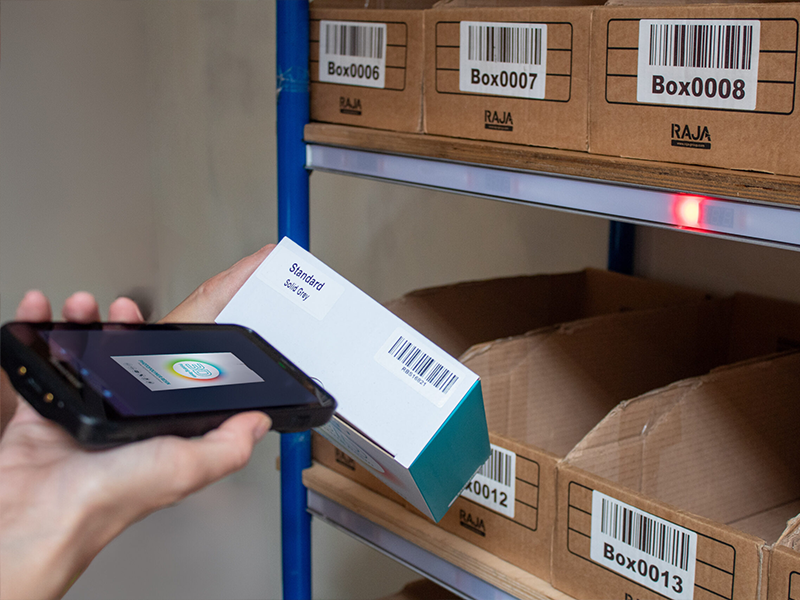 Mitarbeiter scannt ein Produkt mit einem mobilen Gerät vor einem Regal mit beschrifteten Boxen im Lager